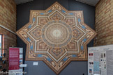 La FRESQUE de la Société Paul Chamoz & Cie de Paray-Le-Monial, Médaille d’or à l’Exposition Universelle de Paris de 1889.
Origine des motifs décoratifs :
« Le style décoratif de la fin du 19ème siècle, que l’on qualifie « d’éclectique s’inspire des styles historiques et reprend, en les mélangeant, les thèmes les plus significatifs des siècles précédents. Ainsi, cette fresque néo-Renaissance dans son ensemble reproduit des motifs décoratifs du 16ème siècle.
Cependant le registre décoratif général de cette fresque s’inspire non des revêtements de sol de l’époque de la Renaissance, mais plutôt de l’ornementation des plafonds :
La partie centrale rappelle les « ciels » en trompe - l’œil de certains décors.
La forme des motifs entourant les « grotesques » reprend celle des plafonds à « caissons » où les bandeaux semblent imiter des moulures dé bois.
Les motifs de coquilles des angles extérieurs reproduisent des décors de lambris du début du 17ème siècle.
L’originalité de cette fresque est dans le choix des coloris, vifs et variés, le bleu notamment, tonalités que l’on retrouve dans la décoration intérieure d’édifices du début du 17ème siècle (chambre bleue du château de Cormatin, en Saône-et-Loire, par exemple).
Enfin, par des dégradés subtils, les couleurs contribuent à donner l’illusion de relief, à créer un trompe - l’œil, tout à fait en accord avec l’esprit de la Renaissance. » Fresque de 40 m2 réalisée en carreaux de grès dessinés par incrustation composée de 700 carreaux de 17,5cm x 17,5cm.