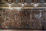 Villa del Principe. Palais d'Andrea Doria.<br> La Salle des Géants. Les tapisseries d'Alexandre Le Grand. La deuxième tapisserie montre un mélange sembleble d’éléments historiques et légendaires et reproduit la Bourgogne du quinzième siècle représentée à travers les costumes, le type d’armes et le style des bâtiments. La partie gauche de l’ouvrage représente la conquête de la ville syrienne de Tyr, en 332 avant Jésus-Christ, qui est ici décrite comme un siège du quinzième siècle. Au centre, un épisode qui appartient à la mythologie sur Alexandre: le souverain macédonien, assis dans une cage décorée de pierres précieuses, tient deux longues tiges au bout desquelles on voit des jambons. Quatre griffons sont enchainés à la cage ; en cherchant d’atteindre les jambons, ils battent les ailes en soulevant leurs corps jusqu’au ciel. Dieu le Père, entouré par des rouges chérubins, suit la scène. Dans la partie droite de la tapisserie, on voit le désir inassouvissable de connaissance qui pousse Alexandre à explorer les abysses de la mer. Le héros se fait plonger au fond de la mer dans une barrique en verre, avec deux flambeaux à la main pour éclaircir l’obscurité des abysses. Enfin, sur l’angle en bas à droite, nous assistons au dernier chapitre du “roman d’Alexandre”, où le souverain macédonien, après avoir conquis tous les peuples du la Terra, arrive au bout du monde et avec ses partisans tue les monstres qui vivent là-bas, représentés comme des êtres à fourrure et sans tête, avec les yeux, le nez et la bouche sur la poitrine.