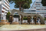 La Grande-Motte. <br> Cette belle place est ornée d’une fontaine dessinée par Jean Balladur lui-même qui dit « … deux présences ajoutent du sens à la figure de cette place. Des jeux d’eaux, jaillissant des lèvres ou des vasques de la fontaine placée en fond de décor y font voir les diableries de l’eau. Le dessin d’un labyrinthe tracé par les marbres rouges et blancs du dallage … rappelle aux autorités qui le dominent … l’importance d’un parcours bien choisi . D’autres voient dans cette eau qui jailli des bouches le symbole des cancans et de la vox populi »