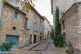 <center>Les maisons de la Grand'Rue.</center> Au Moyen-Age se situait le long de cette rue un quartier consacré à des activités artisanales et commerçantes. L'archéologie du bâti a permis de restituer la physionomie de ce quartier durant le bas Moyen-Age (XIIIème - début XVIème siècle).
Encore présents aujourd'hui, le plus souvent dissimulés dans des maçonneries postérieure, les rez-de-chaussée des maisons de ce quartier présentent de larges arcades de pierre. (ces séries d'arcades répondaient à un souci pratique. Elles permettaient une communication aisée entre les bâtiments donnant sur la rue et des espaces ouverts, installés à l'arrière des maisons). Ces rez-de-chaussée, fonctionnels et largement ouverts sur la rue, étaient conçus pour recevoir les activités artisanales, commerçantes et agricoles du village.
Suite à l'enrichissement de ce quartier s'installent, aux XVIIème et XVIIIème siècles, des petits hôtels particuliers
L'accroissement des échanges au cours du bas Moyen-Age permet à ce quartier de s'enrichir. Les maisons vont donc évoluer vers un nouveau type, moins tourné vers le commerce ou l'artisanat, et devenir des espaces strictement privés. Pour certaines habitations, on retrouve encore leurs aspects des XVIIème et XVIIIème siècles avec des façades qui sont ordonnancées et régulières. On retrouve ici les caractéristiques, des petits hôtels particuliers de village. Le premier étage de ces maisons présente généralement plusieurs grandes fenêtres disposée! symétriquement : il s'agit de l'étage noble où vont se trouver les pièces d'apparat qui seront les plus hautes de plafond ci les plus spacieuses de la maison. Les portes principales sont traitées avec soin, comme le montre l'imposant portail en pierre de taille doté d'un encadrement rectangulaire, visible sur votre droite. Pour ce portail, le motif décoratif est celui d'un bossage (saillie sur la face de parement d'un bloc).